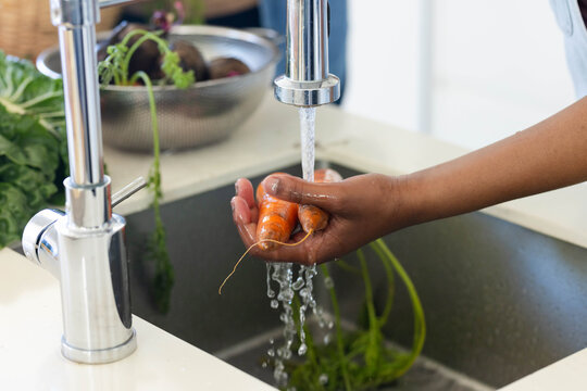 Hand is holding two small carrots under running chrome faucet over steel sink, greens dripping