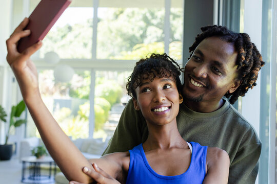 African American couple taking selfie in sunlit living room, woman holding pink smartphone case