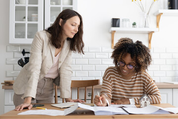 Fototapeta premium business person sitting at table, negotiating. two women are going through papers, discussing project. interior design, architectural bureau. diverse people, African American business woman realtor.