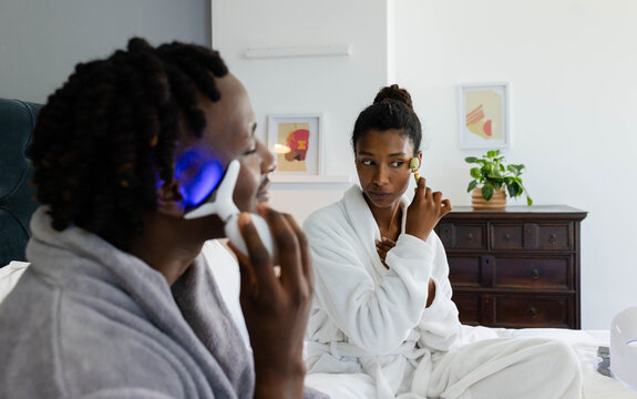 African American couple sitting on bed wearing white robes, using LED device, rolling gold roller