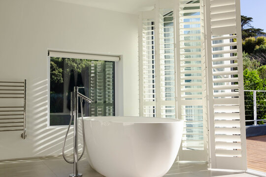 Freestanding oval bathtub is gleaming in bathroom while sunlight is casting shutter shadows to deck