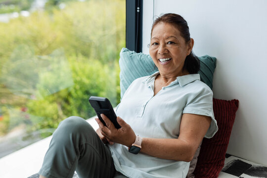 Senior Asian woman sitting on window bench at home holding phone wearing mint polo, smartwatch