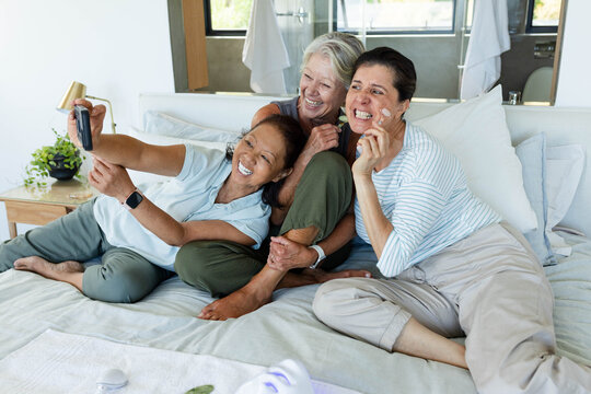 Diverse female friends laughing, posing for selfie on bed with smartphone, facial rollers, pillows