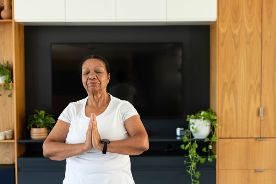 Senior African American woman meditating in living room, wearing white tee  and  smartwatch, TV, pla