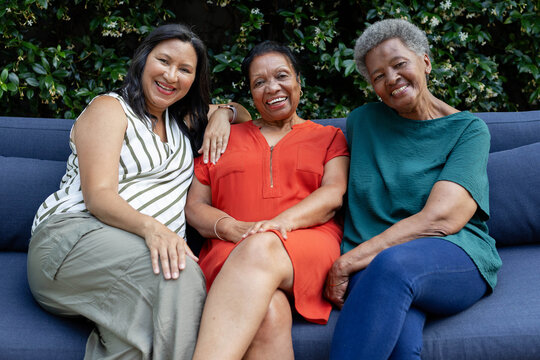 Diverse female friends sitting on navy-blue sofa in garden, smiling and showing bracelet and zipper