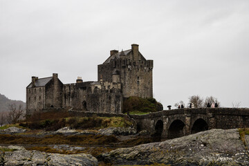 Historic stone castle standing on a rocky landscape under an overcast sky, featuring traditional medieval architecture and a stone bridge leading to the entrance. The scene evokes a dramatic and mood