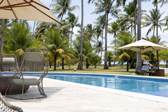 piscina de resort de luxo tropical &agrave; beira-mar (ou beachfront), com &aacute;guas azuis cristalinas, cercada por um gramado verde bem cuidado e dezenas de coqueiros altos (cocos nucifera) balan&ccedil;ando ao vento.
