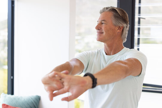 Mature man standing, stretching arms forward in living area by windows, wearing black wristwatch