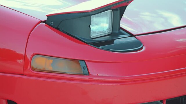 Detailed close-up of a shiny red vintage sports car. Focus on the front bumper, amber turn signal, and closed pop-up headlight. Highlights its classic 90s design and glossy finish.