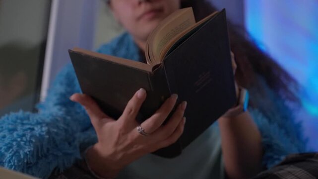 Girl With Dark Cover Reading By Window Light. Portrait Of Girl Immersed In Book Near Softly Lit Window. Young Woman In Cozy Sweater Reading In Quiet Evening By Softly Illuminated Window
