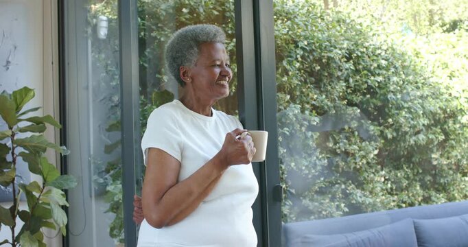 Senior African woman at home by sliding glass door holding tan mug hearing neighbor turning smiling