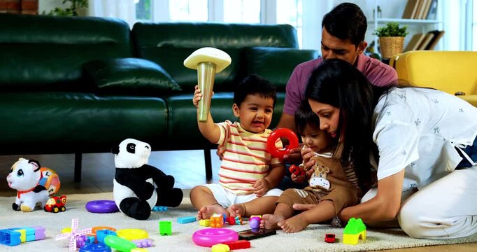 Indian Asian twin infant babies playing with colorful toys while young father and mother sit together on carpet in modern living room at home, capturing joyful family bonding and parenthood moments