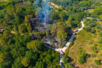 Aerial perspective on a wildfire containment operation. A convoy of fire engines is positioned on a winding dirt road adjacent to the smoldering © AgioiDouloi