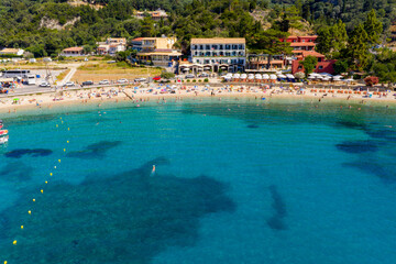 Drone view from the sea looking towards a picturesque Greek beach. Sunbathers relax on the sand under umbrellas while people swim in the incredibly clear, calm, and turquoise Mediterranean water