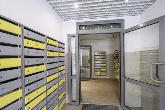 A modern mailroom with rows of gray and yellow mailboxes, glass-paneled doors, tiled floor, recessed ceiling lights, clean minimalist design, leading to another mailbox section