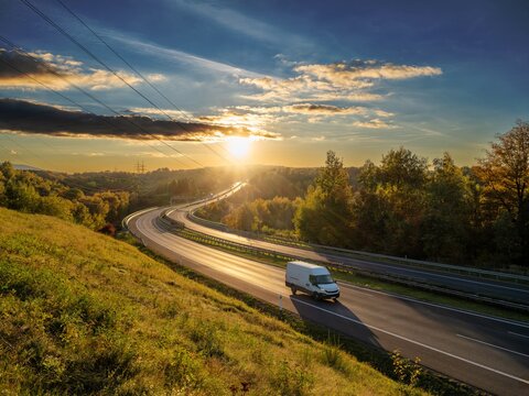 White delivery van driving on a winding highway through an autumn landscape at sunset