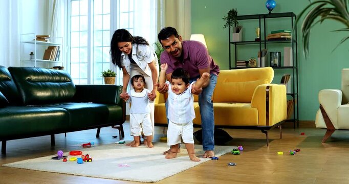 Indian Asian young parents helping twin infant babies walk by holding their hands inside modern home living room, guiding first steps together as smiling babies enjoy playful family bonding moment