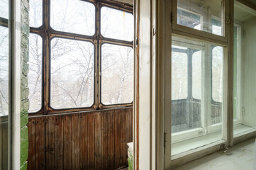 Fototapeta premium A room with large old wooden-framed windows, weathered wooden paneling below, faded green brick wall, overlooking bare trees; balcony visible through glass