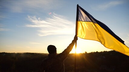 Young soldier of ukrainian army lifted blue-yellow banner in honor of the victory against russian aggression at sunset. Male military in uniform raised a waving flag of Ukraine at countryside