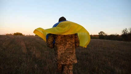 Young male military in uniform jogging with blue-yellow banner on shoulders at meadow. Soldier of ukrainian army running with flag of Ukraine as symbol of victory against russian aggression. Slow mo