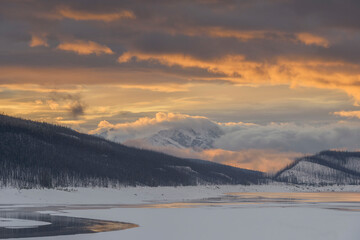 Wide-angle landscape shot of a frozen mountain lake at sunset. Features golden hour clouds, snow-covered peaks, and silhouetted burnt forests. High resolution and balanced composition.