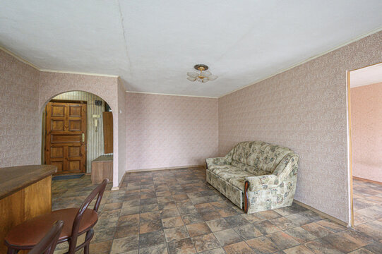 A modest living room with patterned sofa, tiled floor, wooden door, arched doorway, ceiling light fixture, dining chairs, and neutral wallpaper