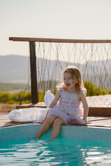 Cheerful girl dangling her legs in the pool. The little girl is very happy and joyful. Mermaid dress. A swim ring with a fish tail lies near the pool. Vertical photo.