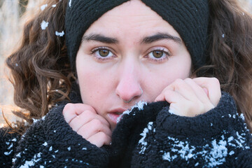 Close-up portrait of a young woman in winter, wrapped in a black fleece jacket and headband, snowflakes on fabric and hair, looking directly into the camera with a calm, introspective expression © ullision