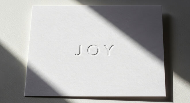 Minimalist white greeting card mockup with elegant embossed JOY text on premium textured paper featuring dramatic diagonal sunlight and shadow for sophisticated stationery design