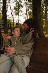 Mom and daughter are sitting on a wooden swing in the park. Funny and lively emotions. The interaction of a mother and a four-year-old girl. Autumn atmosphere. Vertical photo. Mothers Day. 