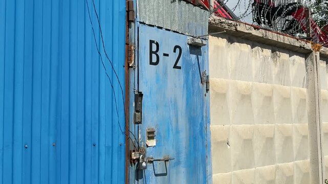 Blue metal door with padlock, weathered gate labeled B against concrete block wall, rusted hinges and chipped paint, overgrown weeds at perimeter of urban construction site security guard.