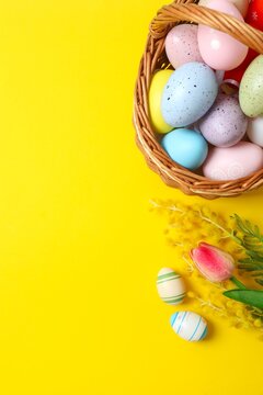 Vertical view of Easter wicker basket full of colorful eggs and spring flowers on bright yellow background, perfect for spring celebrations and greetings.