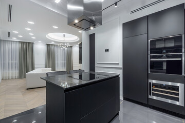A modern kitchen features a black island with sleek surfaces. A living area is visible in the background with curtains and lights on. The scene shows clean lines and a stylish layout
