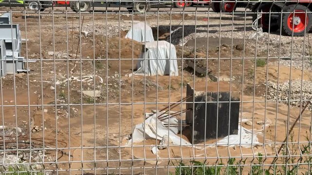 Fenced construction site with sand pile, metal grid fence in foreground, exposed sandy soil, black plastic drainage chamber, wrapped material bundles, scattered pallets, parked trucks
