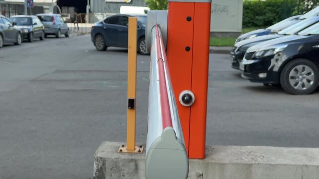 Close-up red parking barrier arm at gated entrance with metal housing, yellow post, asphalt ground, actuator wheel, visible bolts, mild rust, daylight, security access, automatic control.