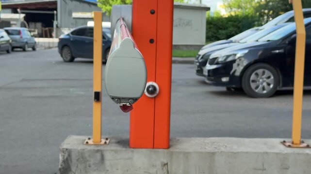 Close-up red parking barrier arm at gated entrance with metal housing, yellow post, asphalt ground, actuator wheel, visible bolts, mild rust, daylight, security access, automatic control.