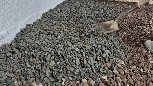 Slow motion close-up of a worker spreading gravel with a shovel along a house foundation.