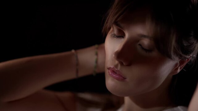 A slow motion portrait of a young woman with her hands behind her head against a black background The lighting is moody and she is posing and looking at the camera conveying a beauty and fashion conce