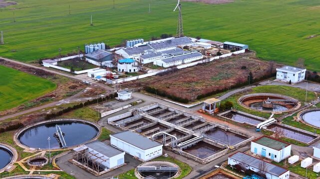 High angle view of municipal wastewater treatment facility complex. Overview of circular and rectangular clarification tanks with surrounding buildings and green fields.