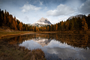 Lake antorno reflecting tre cime alpine mountains, creating a mirror image with vibrant autumn larch forests and cloudy sky
