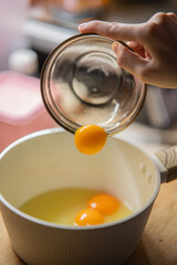 Hand pouring fresh egg yolk from glass bowl into a white cooking pot with other eggs, preparing food