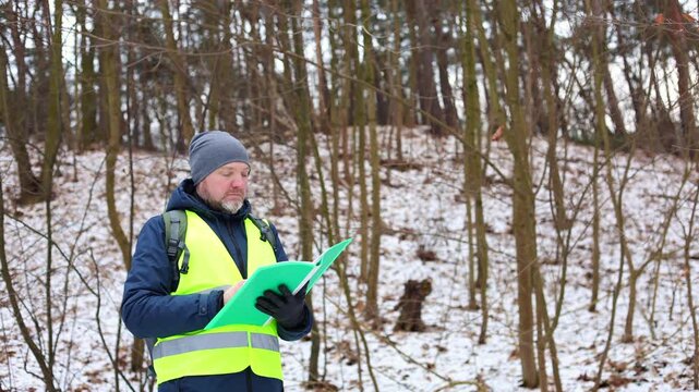Environmentalist forest inspector in a safety vest evaluating the condition of trees in a snowy winter woodland. Specialist is conducting ecological monitoring, checking for pests or disease.