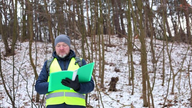 Environmentalist forest inspector in a safety vest evaluating the condition of trees in a snowy winter woodland. Specialist is conducting ecological monitoring, checking for pests or disease.