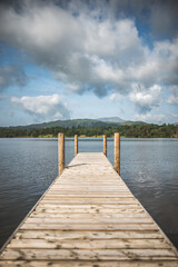 Obraz premium Wooden pier extending into a calm lake with forested hills and cloudy sky