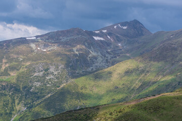 Obraz premium Sunlit alpine mountain slopes with patches of snow under dramatic cloudy sky