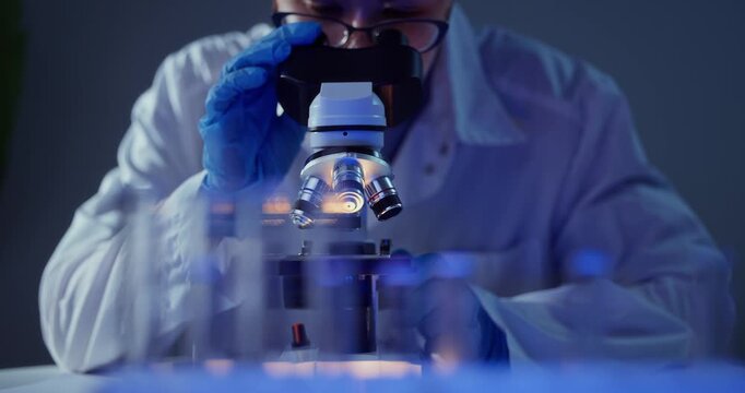 Scientist adjusts microscope objectives and focuses under blue laboratory lighting, gloved hands rotating lenses and peering closely at samples.