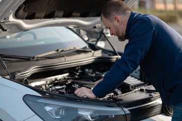 Male mechanic inspects car engine under the hood, wearing a blue jacket, with a focus on automotive repair in an outdoor setting © Kateryna Muzhevska