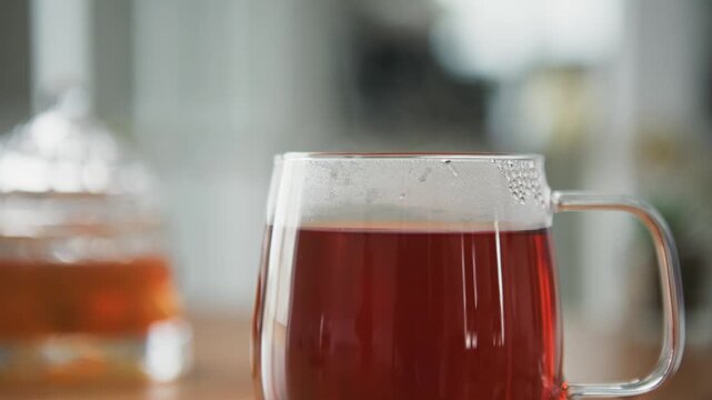 Detailed macro: transparent glass cup of hot tea stands on wooden table in bright living room with morning sunlight. Thick steam rises elegantly, evoking cozy breakfast, warmth and daily relaxation 