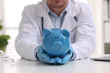 Doctor with piggy bank at white table indoors, closeup