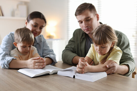 Family with Bible praying together at wooden table indoors